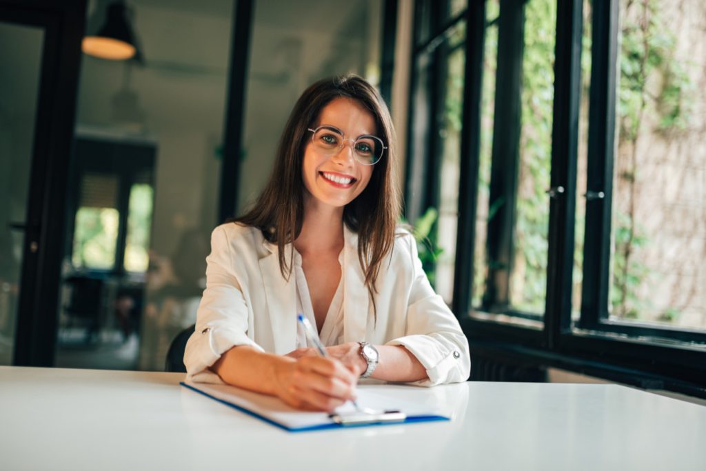 Junge Frau mit Brille sitzt an einem Tisch und schreibt auf einem Blatt Papier